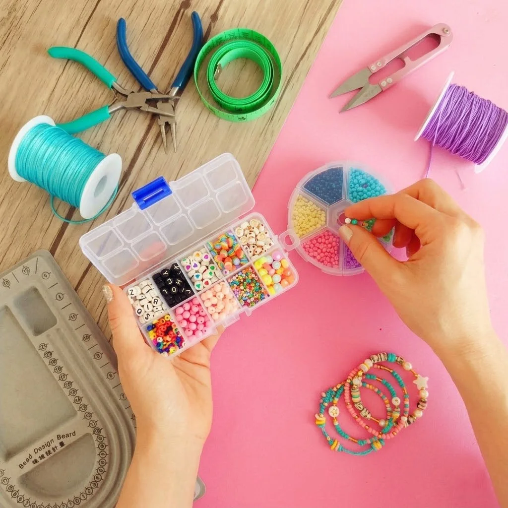 Una composición fotorrealista en formato cuadrado (1:1) que combina elementos clave para aprender bisutería como negocio. En el centro, unas manos profesionales organizan cuentas de colores de un organizador de plástico. El diseño incluye herramientas profesionales de bisutería (pinzas, cinta métrica, tijeras rosa), un tablero de diseño de cuentas y spools de hilo sobre superficies contrastantes de madera y rosa pastel, ilustrando el proceso de creación y organización para un emprendimiento artesanal.
