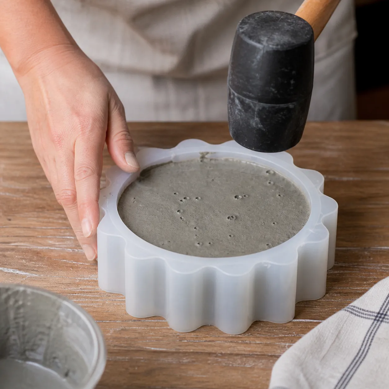Una mano artesana golpeando suavemente un molde de silicona lleno de concreto fresco sobre una mesa de madera, con burbujas de aire subiendo a la superficie de la mezcla gris.