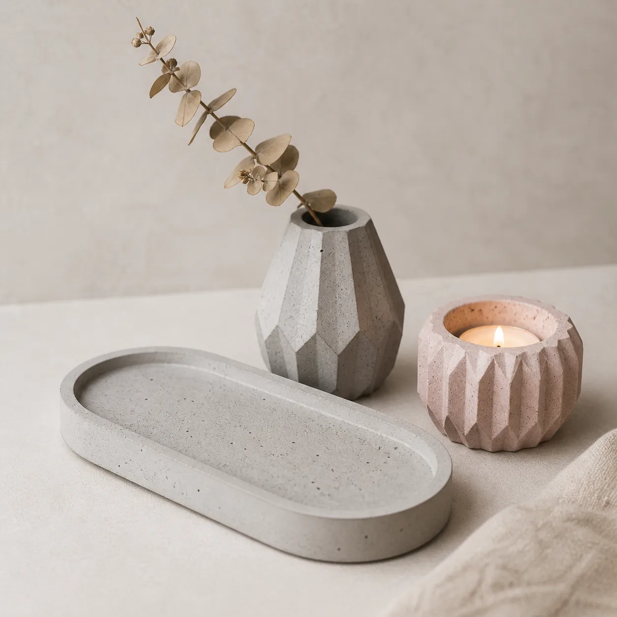 Una composición elegante de piezas terminadas de concreto en tonos pastel y gris natural: una bandeja ovalada, un portavelas y un pequeño jarrón con una rama seca sobre un fondo neutro