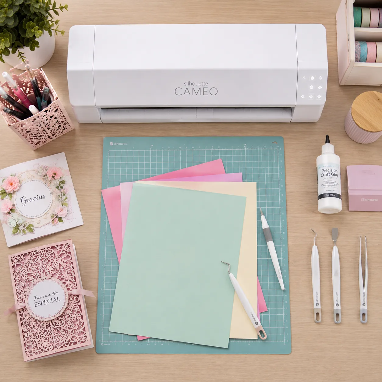 Vista cenital de un escritorio de trabajo limpio con un plotter Silhouette Cameo blanco, tapete de corte con cartulinas de colores, herramientas de depilado, pinzas de precisión y pegamento de punta fina.