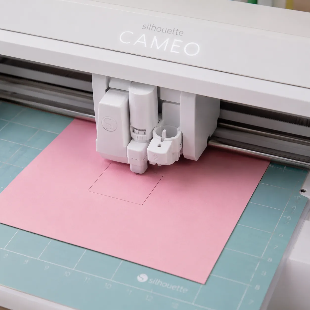 Primer plano de una máquina Silhouette Cameo realizando una pequeña prueba de corte cuadrada en una cartulina rosa pastel sobre el tapete de sujeción.