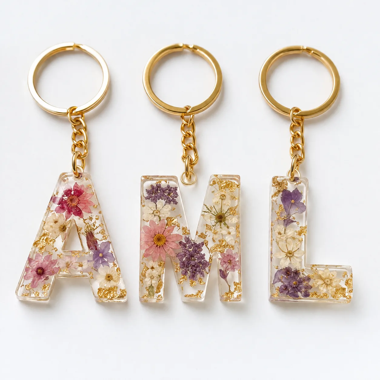 Tres llaveros de letras en resina epóxica con hojas de oro y flores secas encapsuladas, colocados de forma ordenada sobre una superficie blanca.