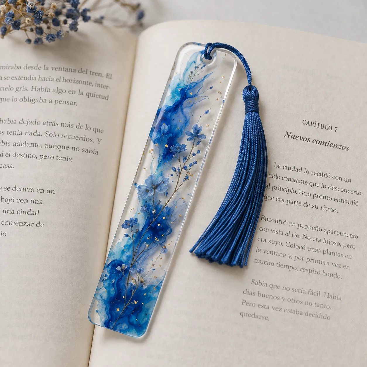 Un marcapáginas de resina transparente con detalles de pintura acrílica azul y una borla de hilo de seda, colocado sobre un libro abierto.