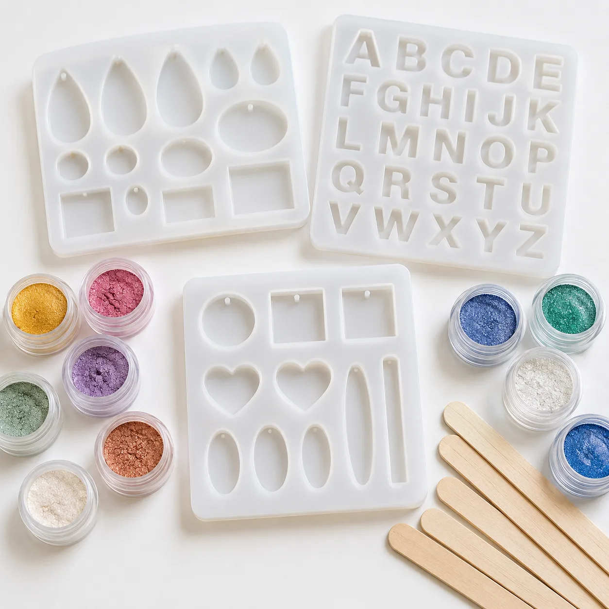 Tres moldes de silicona flexible para bisutería y letras, rodeados de pequeños frascos con polvos de mica brillantes y palitos mezcladores sobre un fondo blanco.